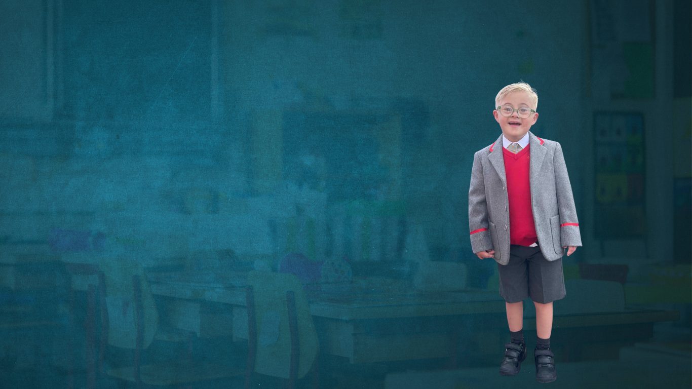 A young child with down syndrome wearing glasses and in a very smart school uniform wearing a blazer and shorts and tie. The young child is smiling into the camera and standing with his hands by his side