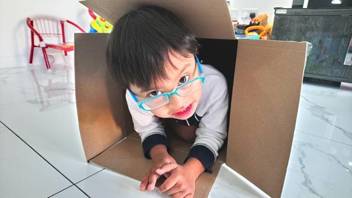 Child with down syndrome climbing out of a cardbox box wearing glasses