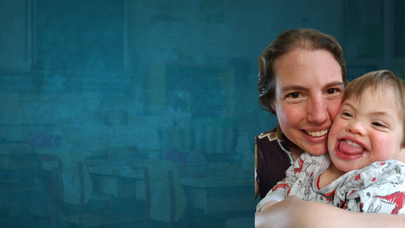 Mum holding her son from behind and smiling for the camera, the young boy is looking to the left, with a big grin and his tongue sticking out