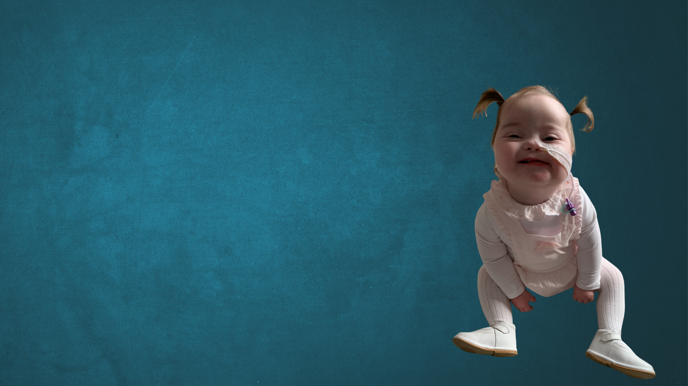 Child with down syndrome sitting on the floor with her feet in front smiling with head up and with pigtails. She's showing an NG Tube coming out of her nose