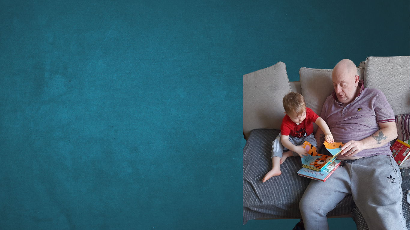 Man sat on the couch holding onto a book that his grandchild is also holding onto and looking at the pages intently