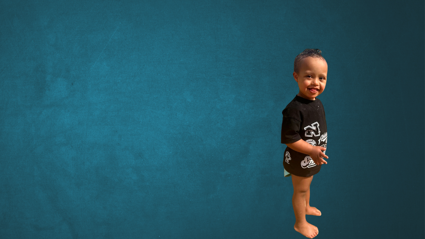 Child with down syndrome standing with his side facing the camera and his head turned smiling with a cheeky grin