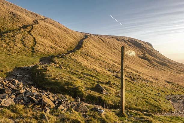 Pen-y-ghent mountain from below with a national trust hiking sign in the image and a stunning day with sun rays glowing off the mountain