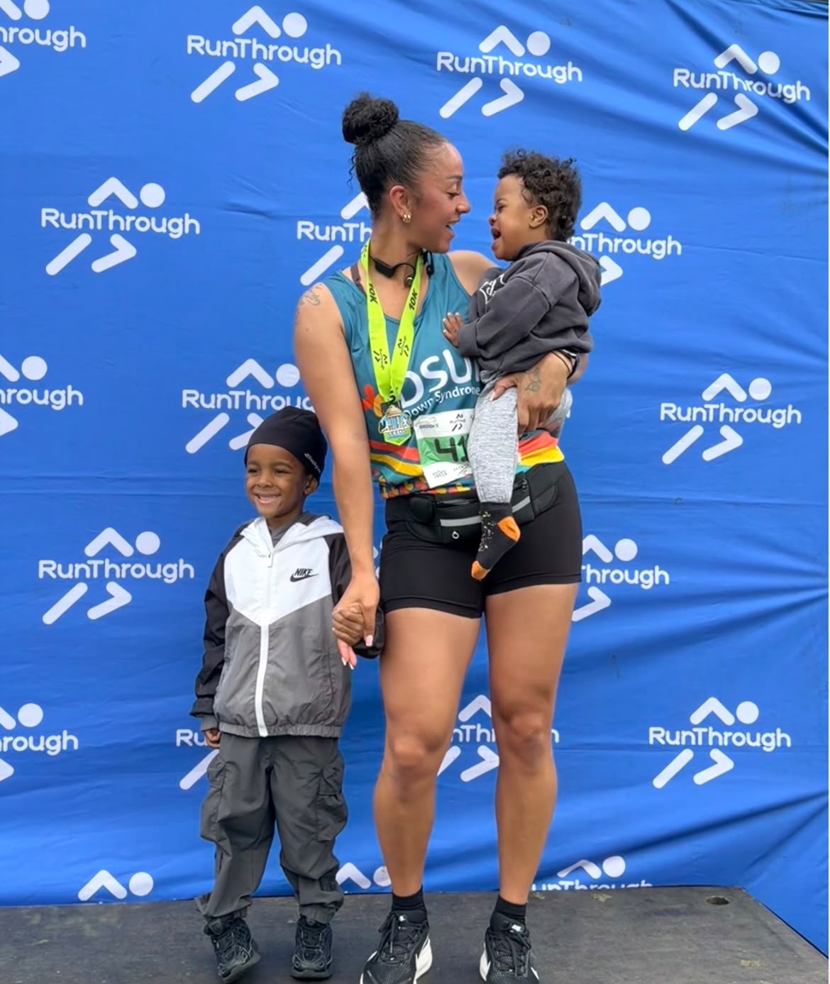 Faith lacey holding her kids hand in one hand and her baby in her arm of the other hand, posing for a picture in her DSUK running vest as she completed a race