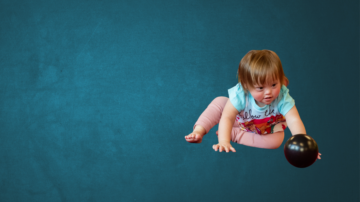 A toddler with Down syndrome reaches for a ball while crawling on a red mat, with soft play skittles in the background, showing gross motor play and physical development in action.