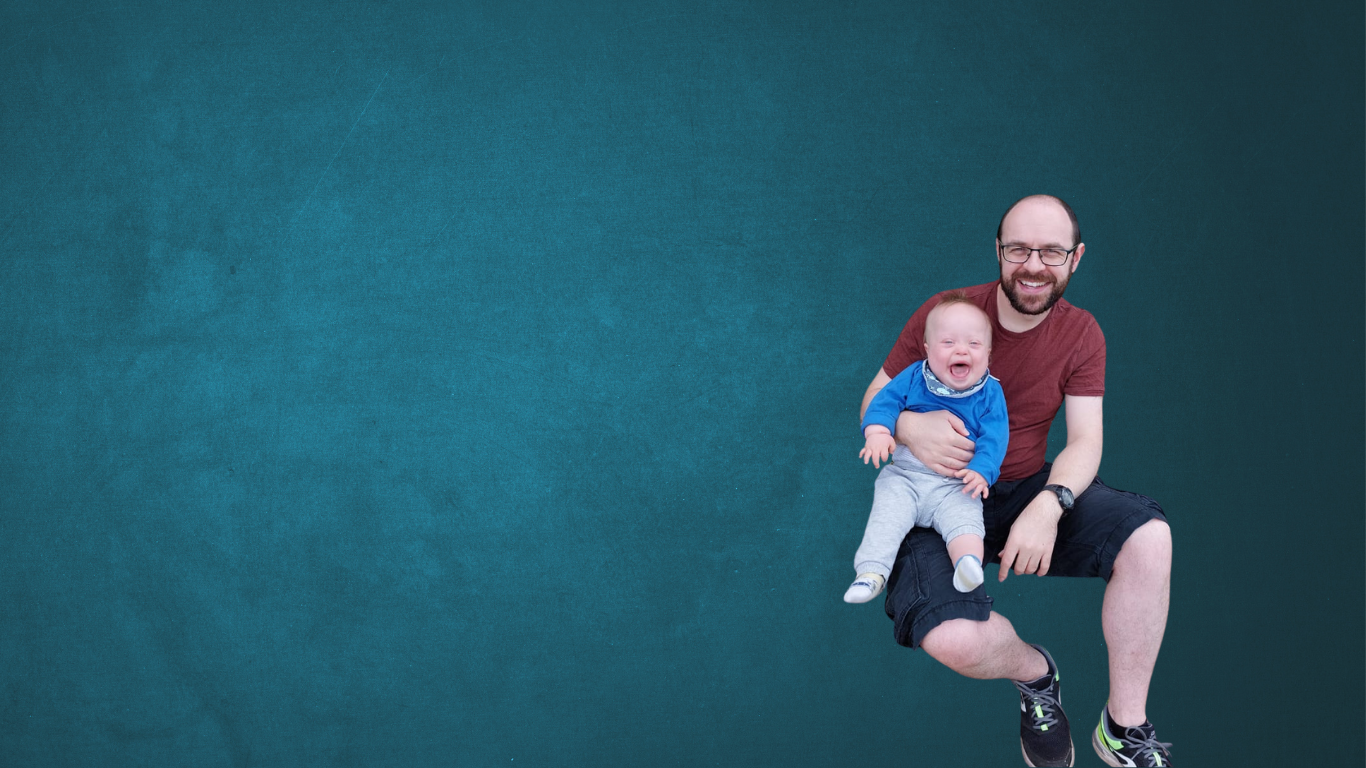 A father smiles while holding his laughing baby with Down syndrome on his lap, set against a teal background.