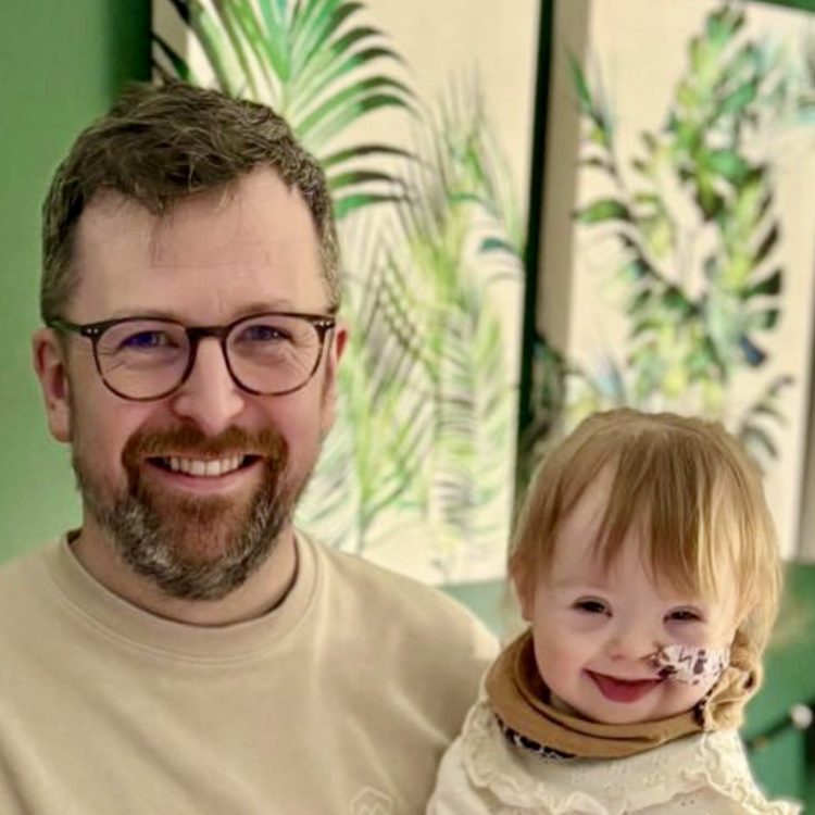 David is posing with Norah in this photograph. Norah has an infectious grin and is wearing a scarf. She also has an NG tube
